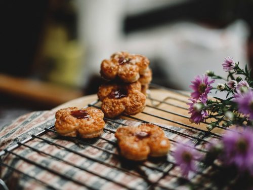 Blütenförmige Gebäckstücke mit Marmelade auf einem Kuchengitter, umgeben von lila Blumen, ideal für Backliebhaber und Feinschmecker.