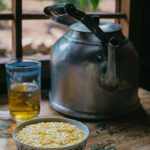Eine Schüssel mit gelbem Blumenkohlsuppe steht neben einer Teekanne und einem Glas Tee auf einem Tisch.