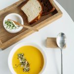 Schüssel mit Kürbissuppe, frischen Kräutern, Joghurt und zwei Scheiben Brot auf einem Holzbrett.