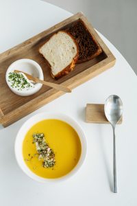 Schüssel mit Kürbissuppe, frischen Kräutern, Joghurt und zwei Scheiben Brot auf einem Holzbrett.